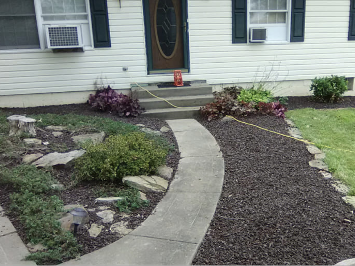 A front yard with a curved concrete walkway leading to steps at the house entrance, landscaped with bushes, plants, rocks, and mulch, with two deck chairs near the door.