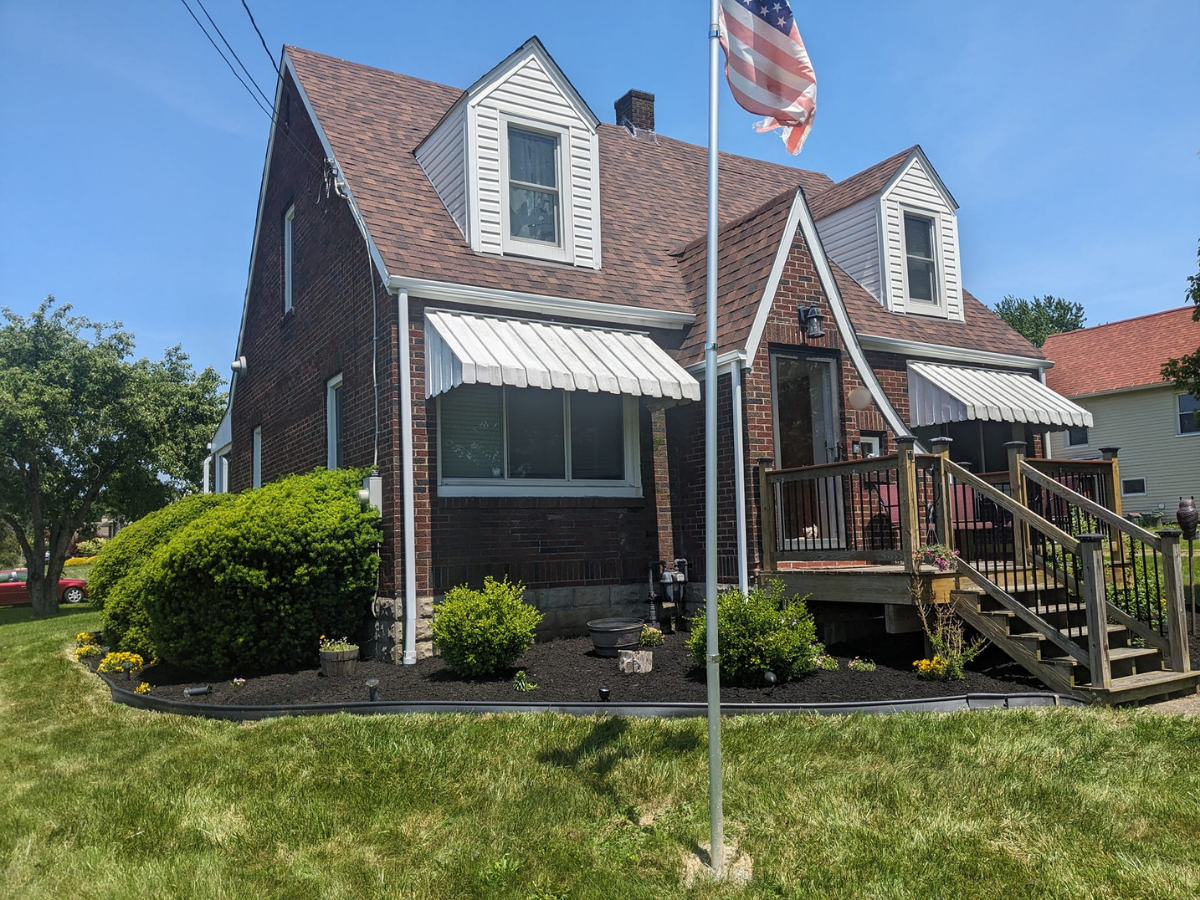 Front view of a brick house with dormer windows, white window awnings, a wooden porch with stairs, and an American flag on a flagpole in the yard, under a clear blue sky.