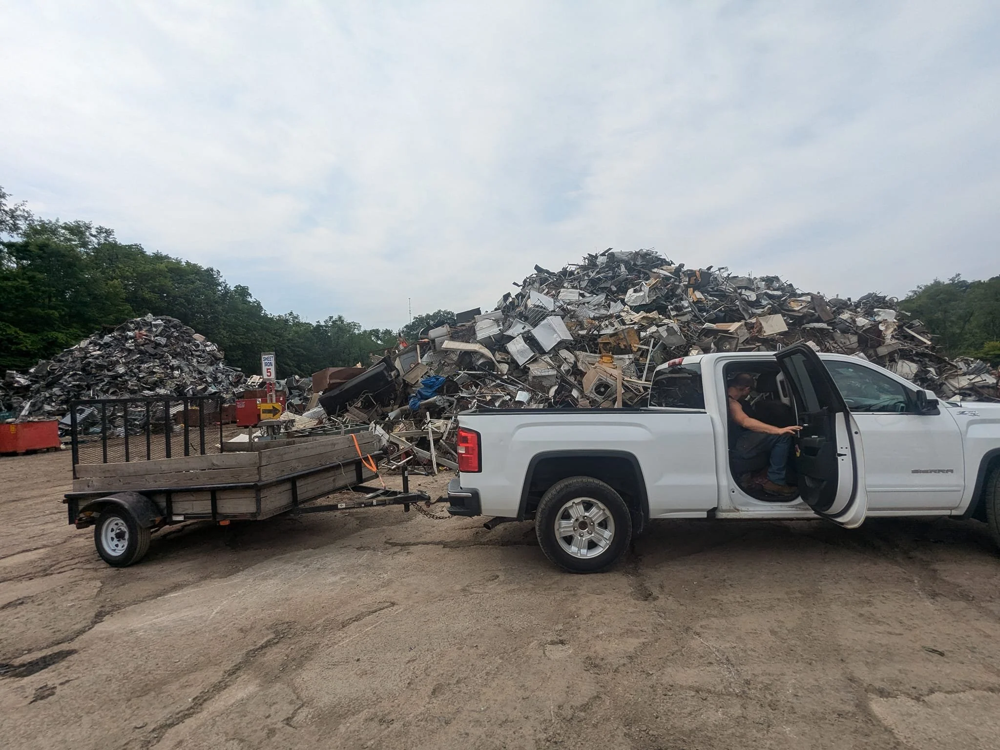 White pickup truck pulling a trailer loaded with scrap metal and appliances, parked at a salvage yard with large piles of scrap metal in the background under cloudy sky.