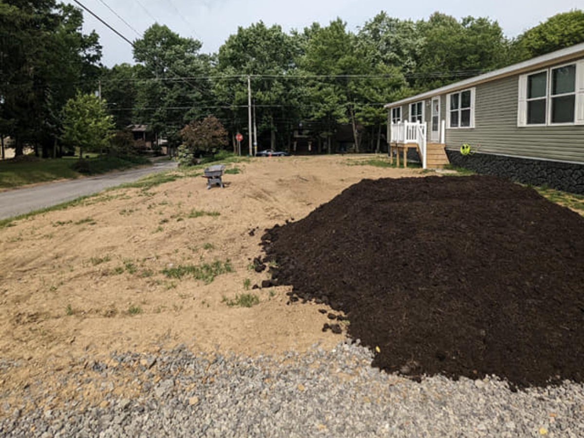 A yard with a pile of dark soil, a patch of gravel, and a mostly bare area of dirt with some grass, next to a house with gray siding and white trim, during daytime.