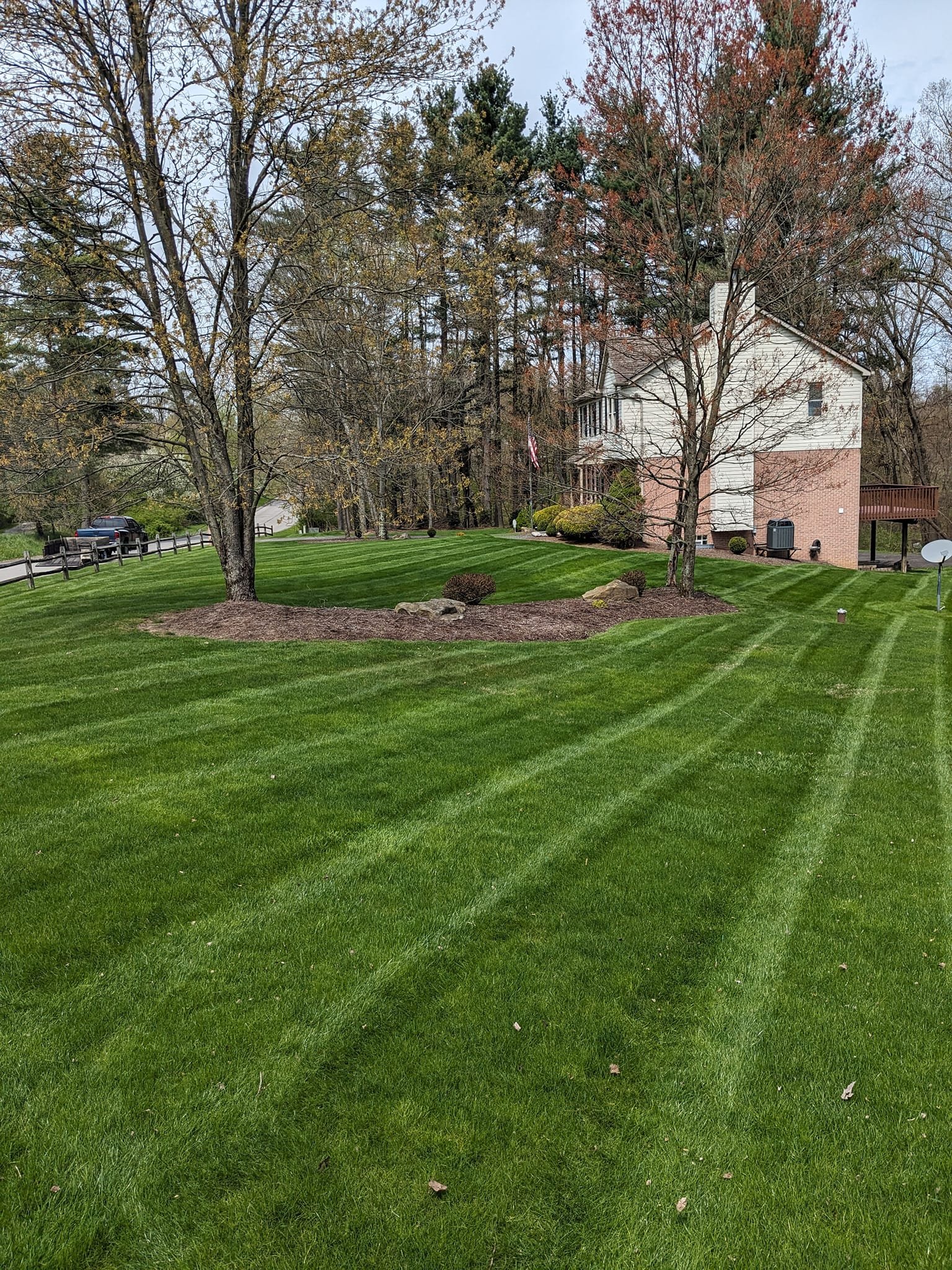 A house with white siding and a brick foundation, surrounded by a well-manicured lawn with striped grass, trees with some leaves, and a cloudy sky.