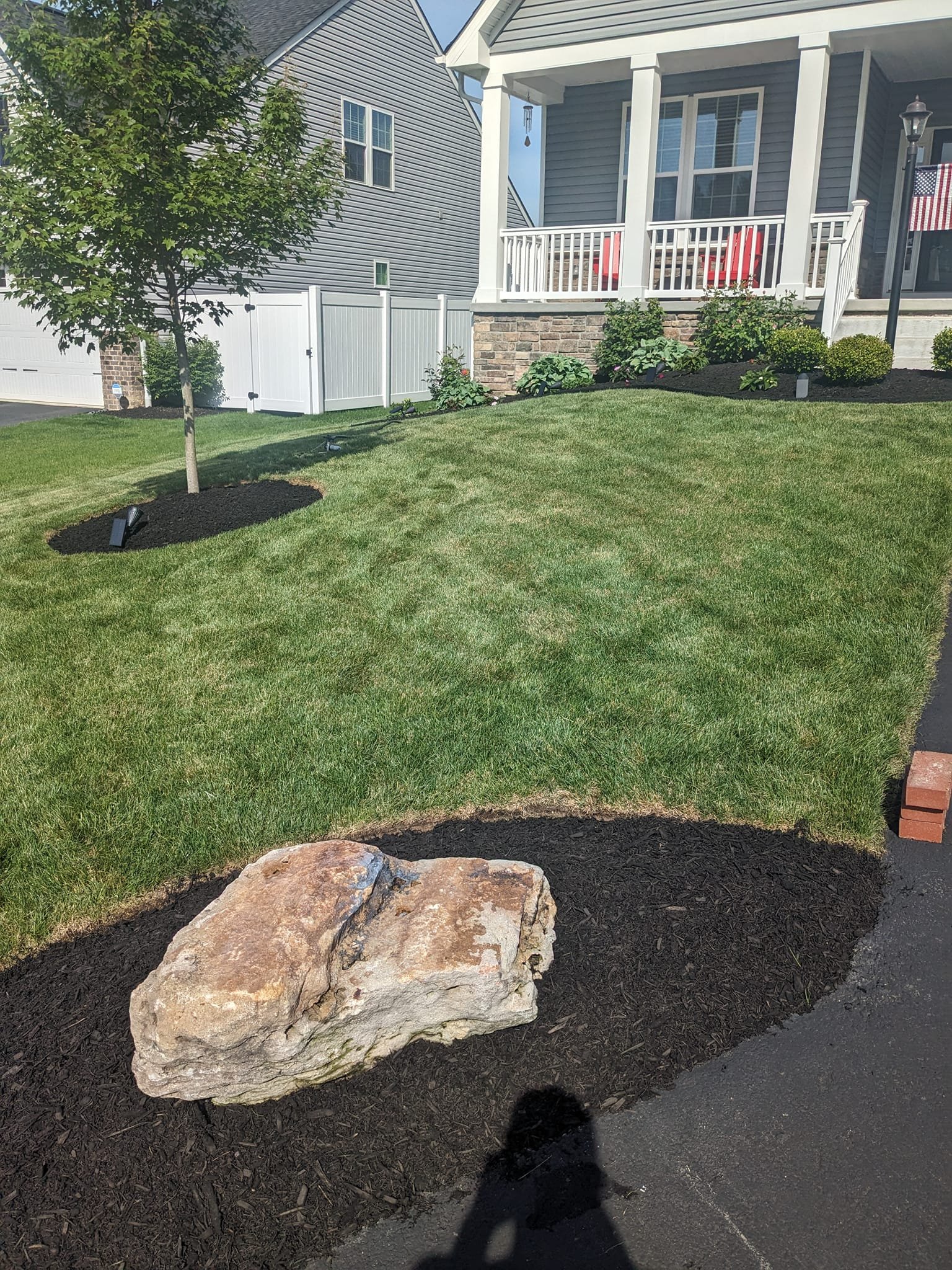 A front yard with manicured green grass, a young tree with mulch around it, a large rock in front, and fresh mulch beds with plants near a house with a porch and white fence.