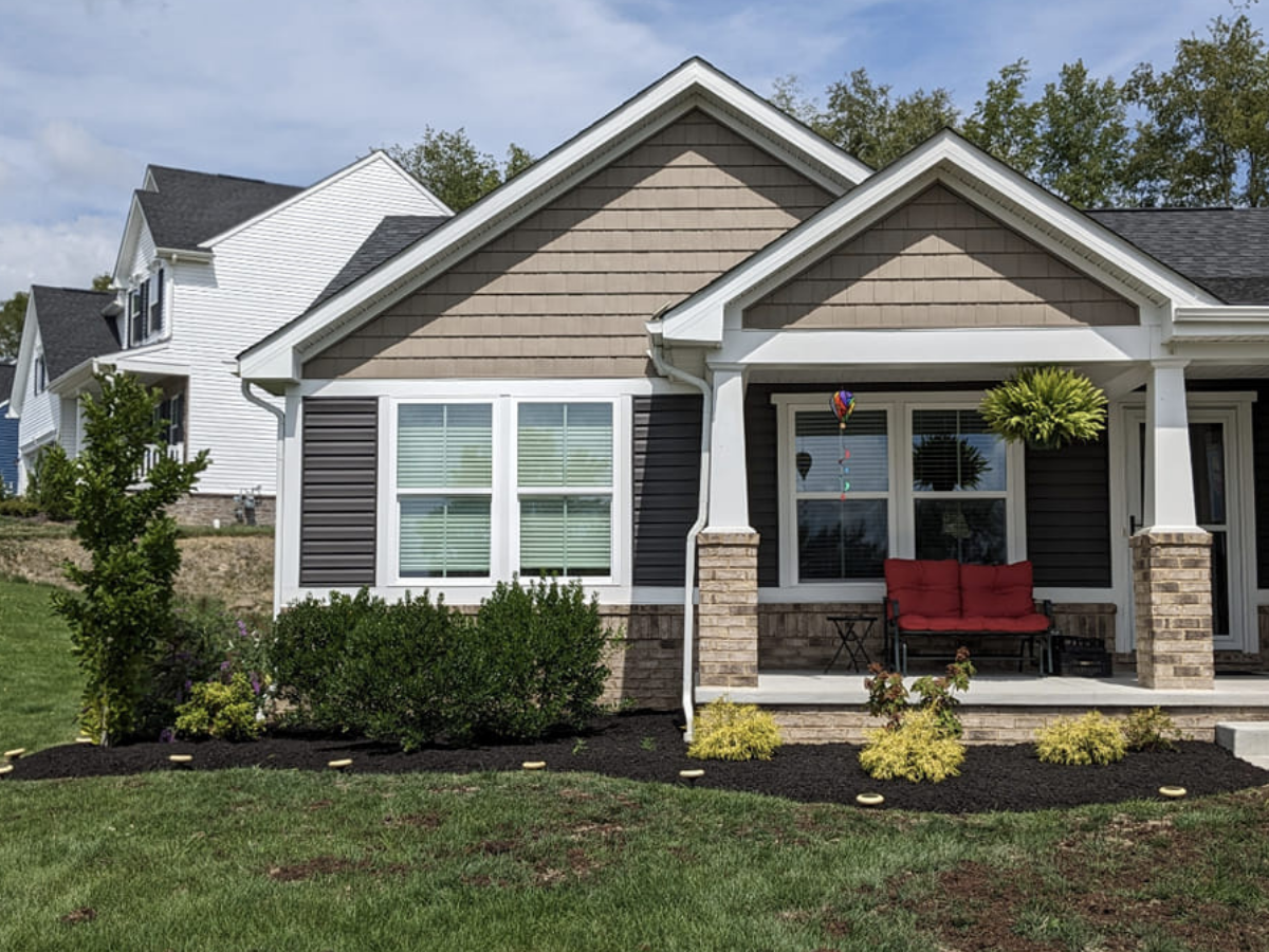 Front view of a modern house with a porch, a red outdoor sofa, hanging plants, and a landscaped yard with shrubs and yellow plants.