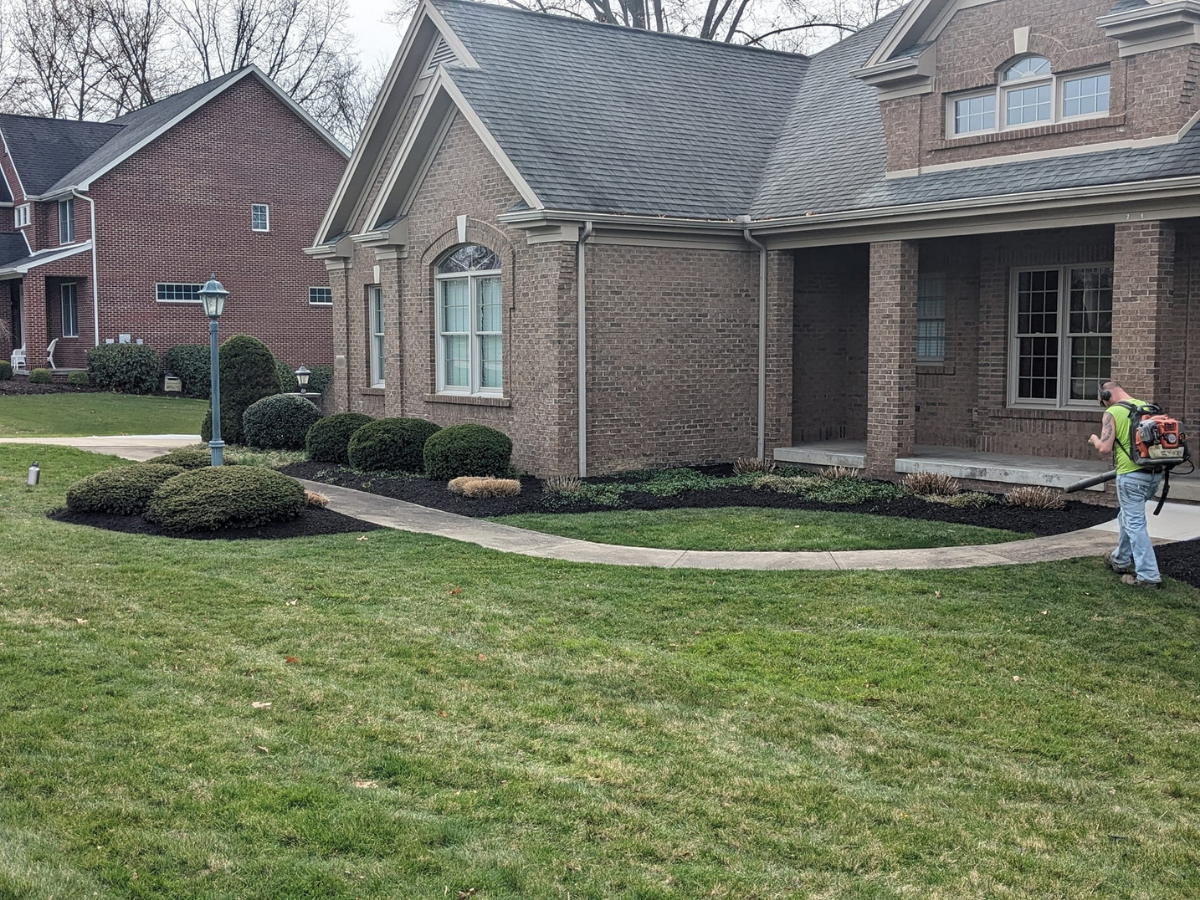 Man operating a leaf blower in front of a brick house with a landscaped yard and sidewalk.