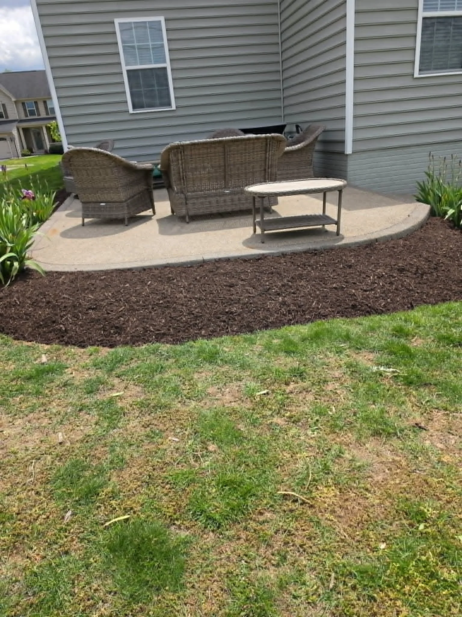 A backyard patio area with wicker outdoor furniture, including a sofa and chairs, on a concrete slab. There are green plants and flowers along the edge of the patio, and a house with gray siding and white trim in the background.