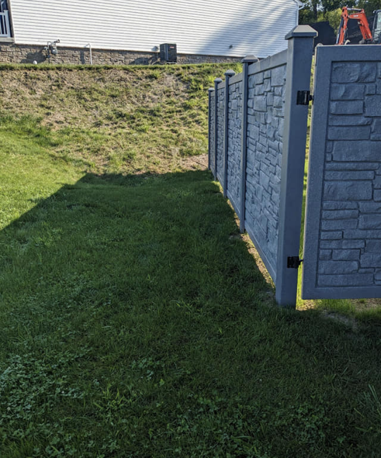 A gray stone textured fence with a gate, running along a grassy area and a sloped dirt hillside in the background.
