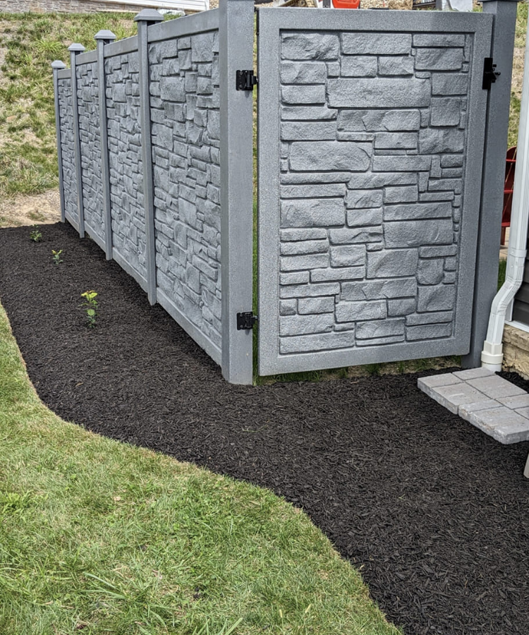 A newly installed gray fence with stone pattern panels, opening to a small garden area with a few young plants, surrounded by black mulch and grass.