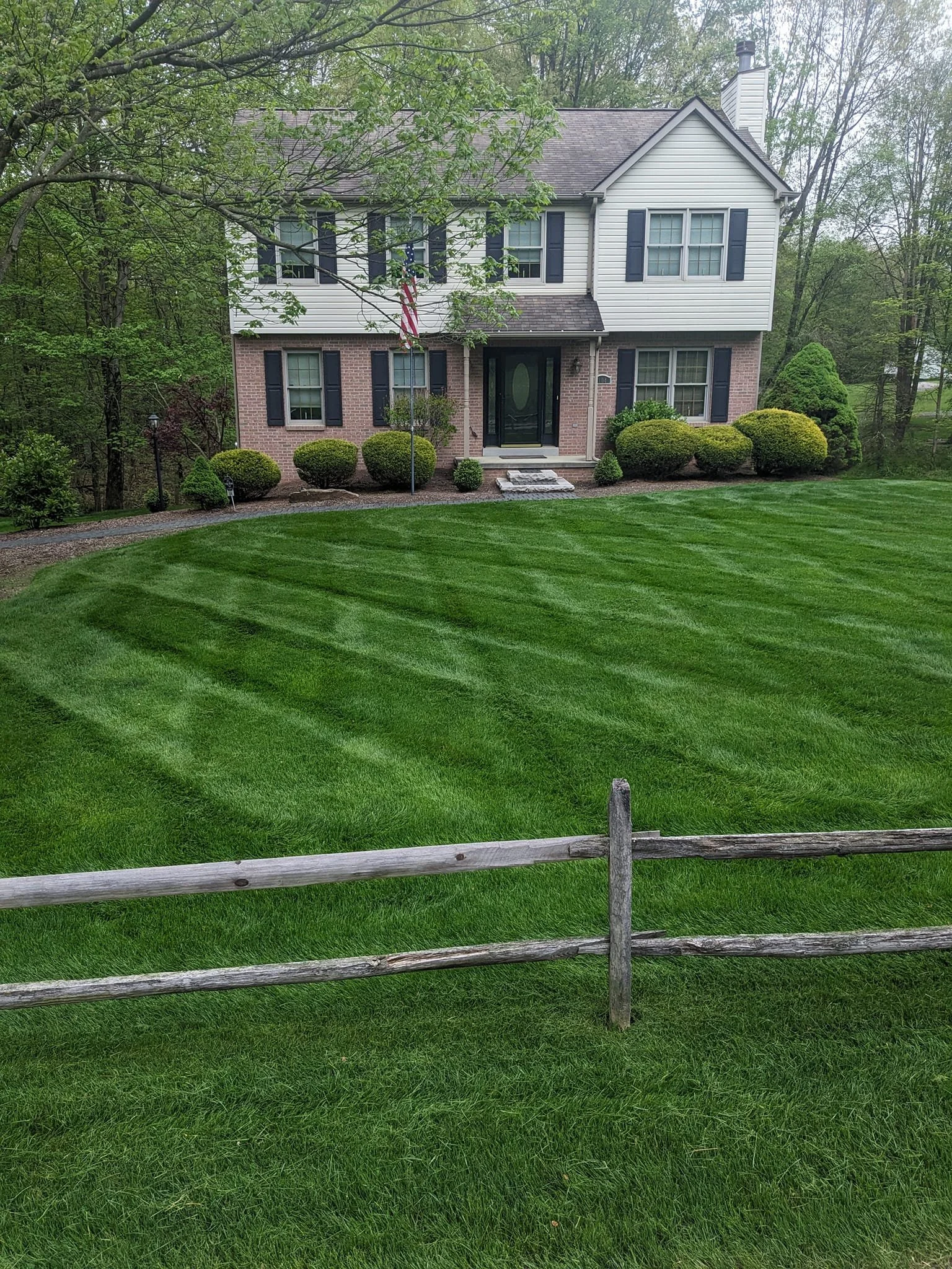 A two-story house with white siding and brick facade on the lower level, black shutters on the windows, a front door with stairs, surrounded by green bushes and a well-maintained lawn, with a wooden fence in the foreground and a wooded area in the ba