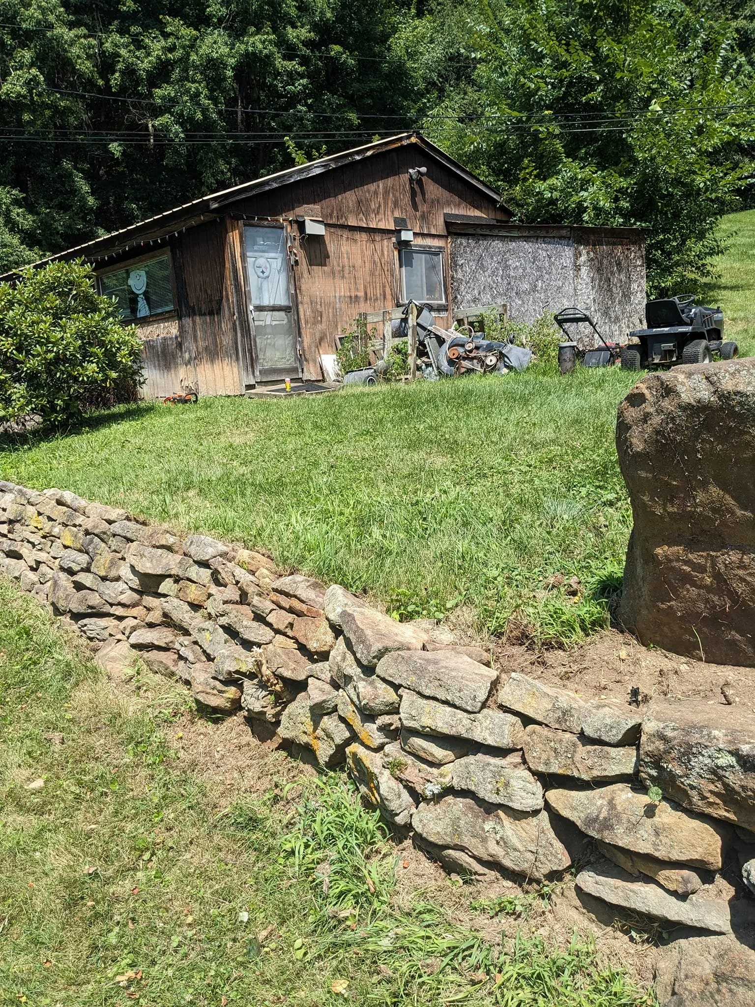 A small wooden shed with a sloped roof, surrounded by green grass and trees. There is a pile of rocks arranged in a line in the foreground, and various outdoor objects such as a lawnmower, motorcycle, and plants are scattered around the shed.