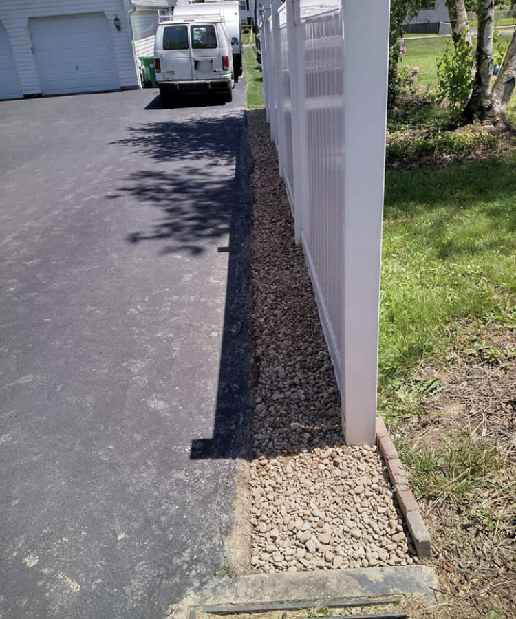 A driveway next to a white fence with gravel at the base, and a parked white van in the background.
