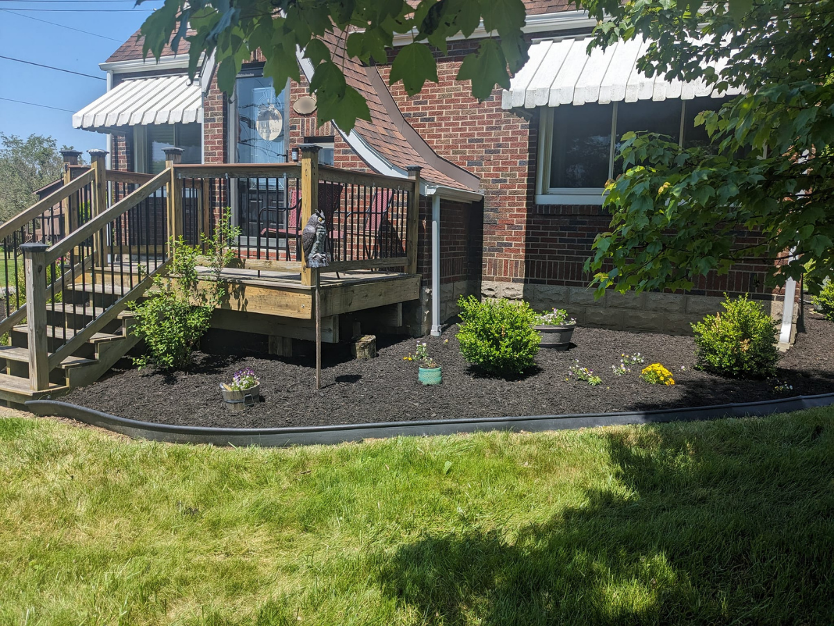 A brick house with a small wooden deck, black railing, and white awnings. There are some plants, shrubs, and flowers in a landscaped garden bed in the foreground, with a grassy lawn and trees around.