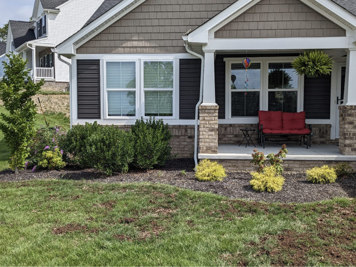 The front yard of a house with a porch that has a red outdoor bench, windows with white blinds, a hanging lantern, and landscaped garden with bushes and small yellow plants.