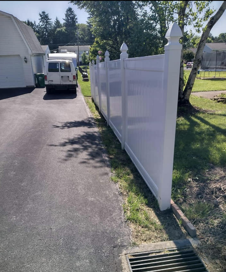 White vinyl privacy fence extending along a paved driveway next to a grassy yard, with a white van parked nearby and tall trees in the background on a sunny day.