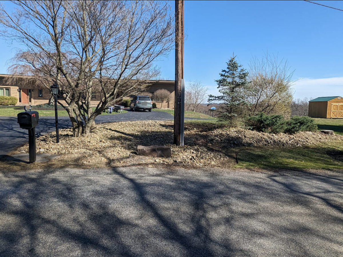 A house with a driveway, a parked vehicle, and a mailbox. There are trees, a utility pole, and a shed in the yard, with a clear blue sky overhead.