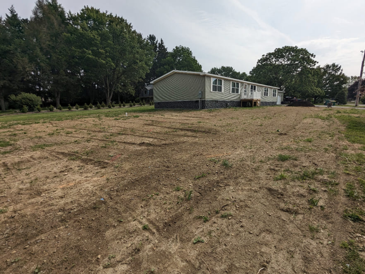 A house with a white exterior and multiple windows, with a raised porch, is situated behind a dirt yard with sections prepared for planting, surrounded by trees and grass on a cloudy day.