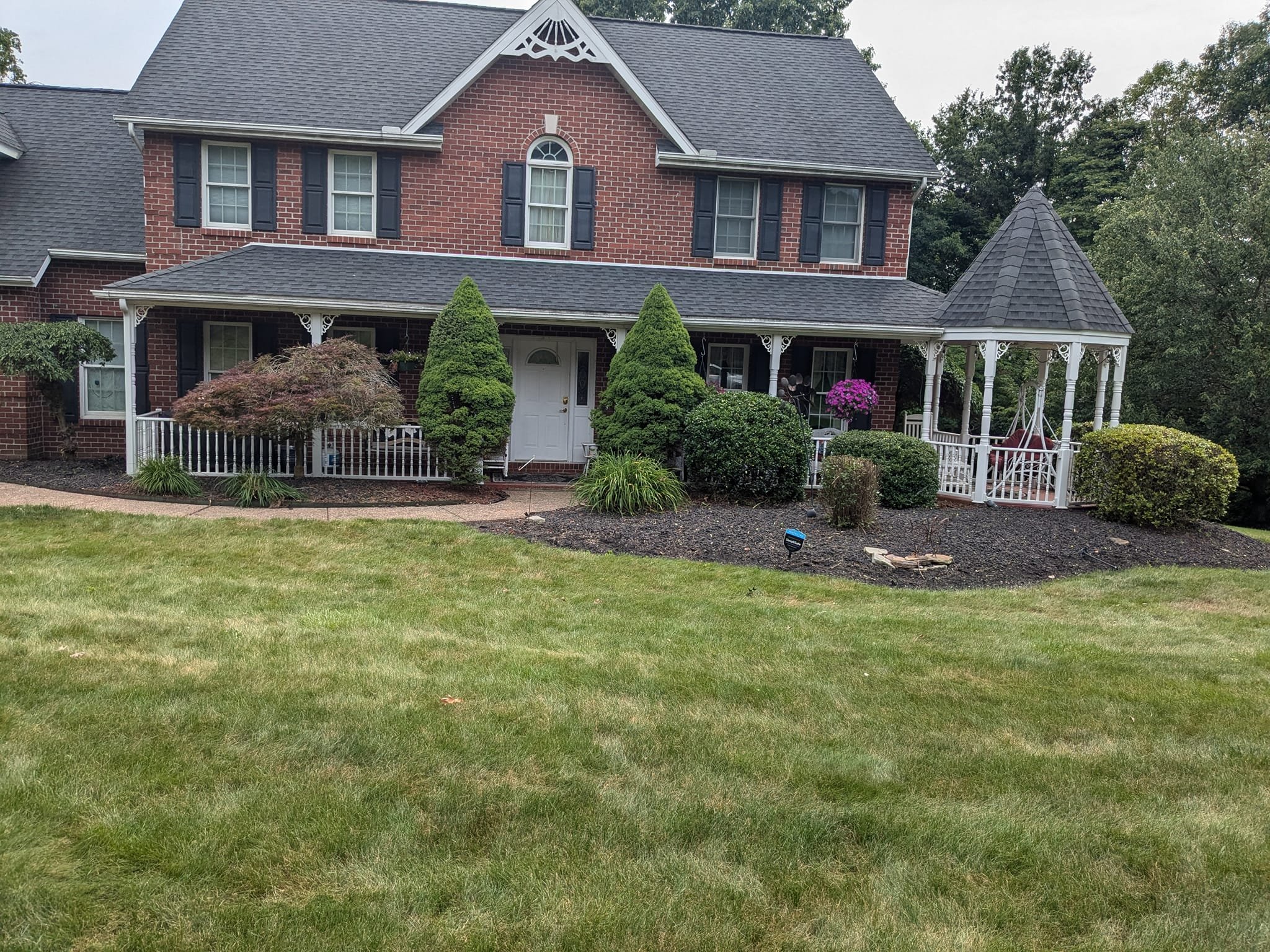 A large brick house with black shutters on the windows and a gabled roof. There is a front porch with decorative white trim, bushes and trees in the front yard, and a small white gazebo with a swing on the right side of the house.