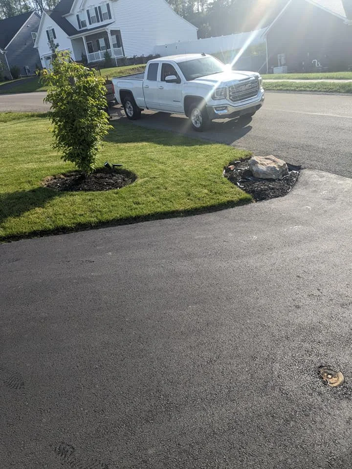 White pickup truck parked on the street near a small tree and a large rock, with a grassy yard and houses in the background.