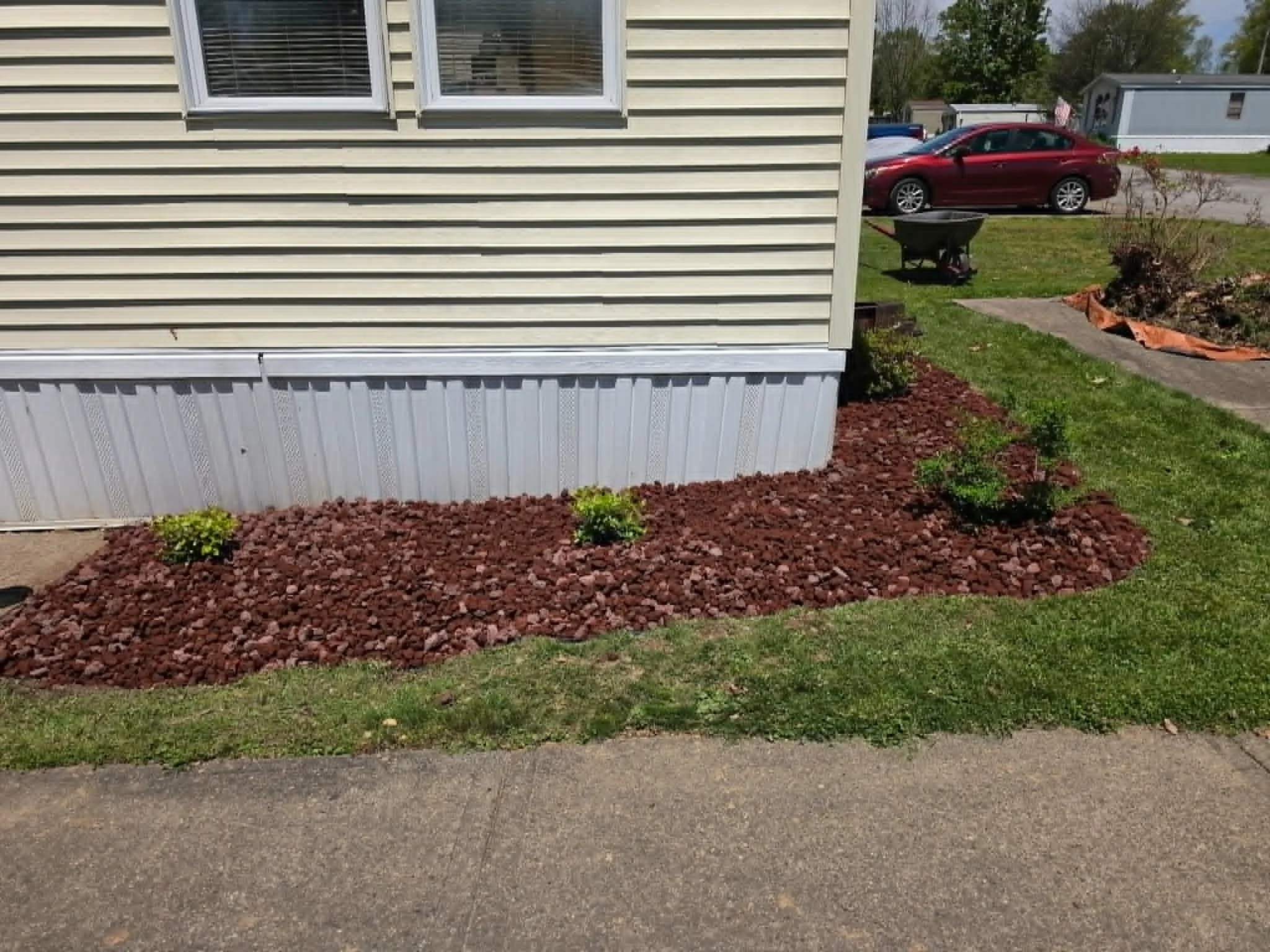 View of a house with beige siding, two attached windows with white blinds, a small garden bed with red mulch and small green plants, a grassy yard, a concrete sidewalk, and parked cars including a red car and a gray car in the driveway. In the backgr