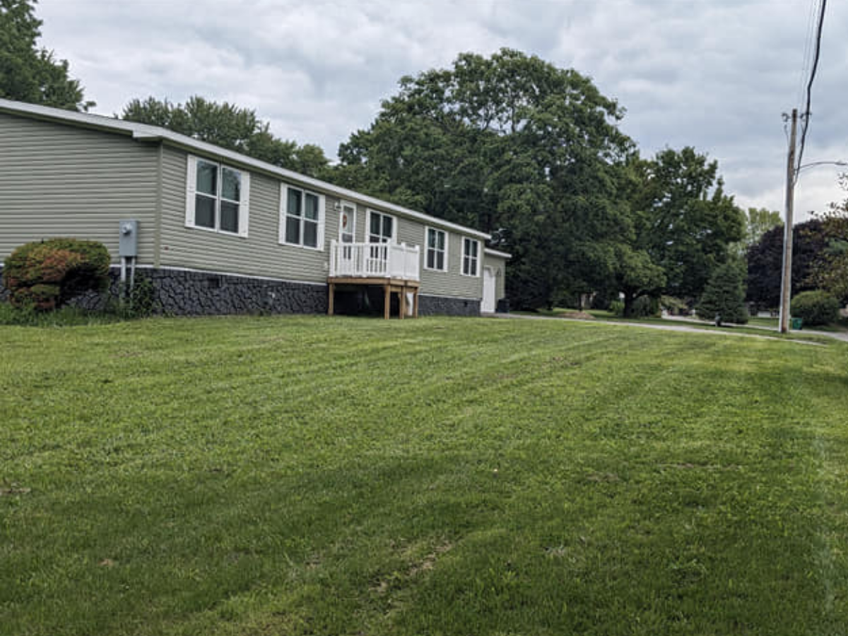 A single-story house with a gray exterior, white trim, and a raised deck with stairs, set on a grassy lawn with trees in the background and power lines to the right.