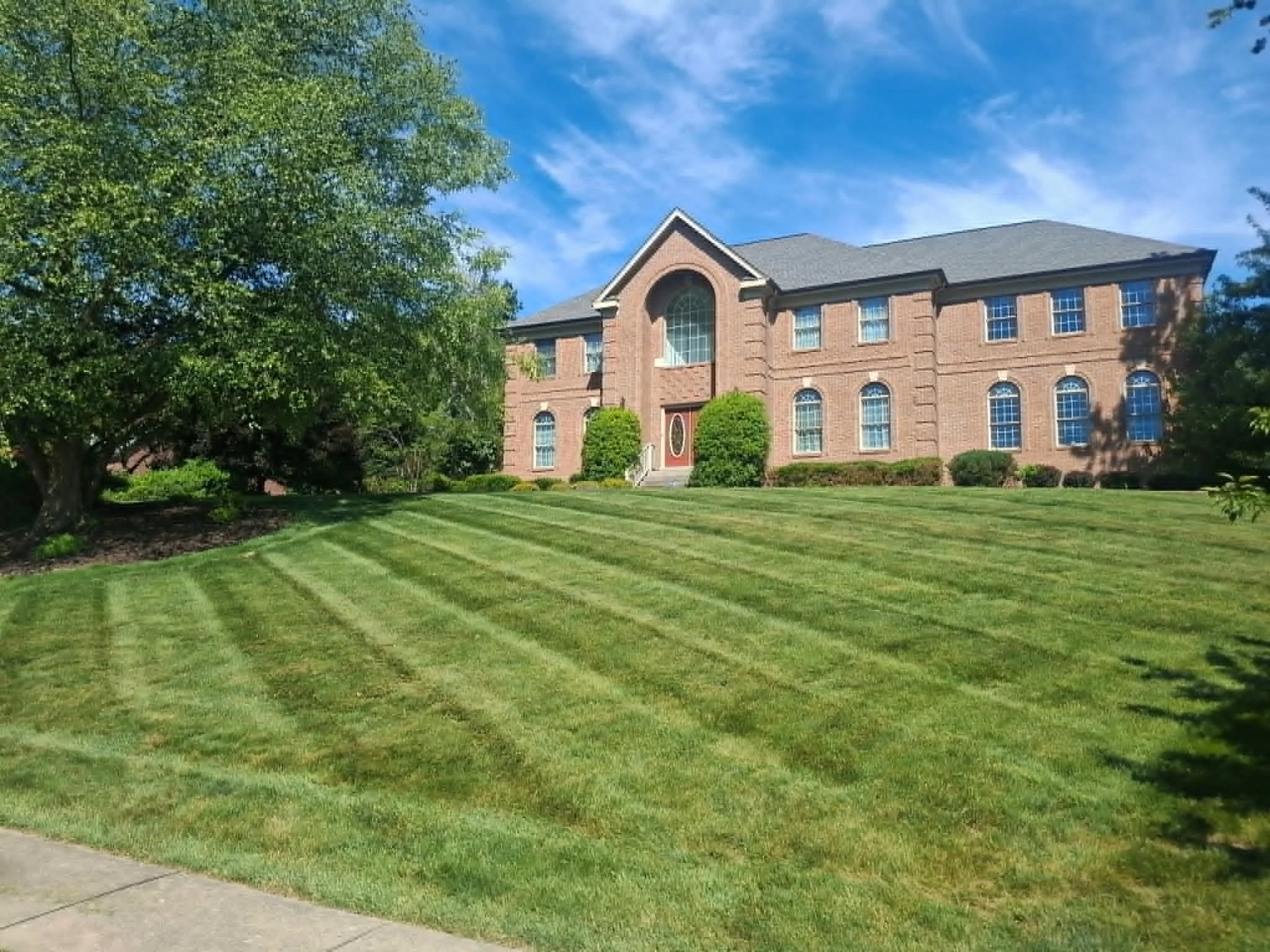 A large brick house with multiple windows and a gray shingled roof, surrounded by a well-maintained lawn and trees under a mostly clear blue sky.