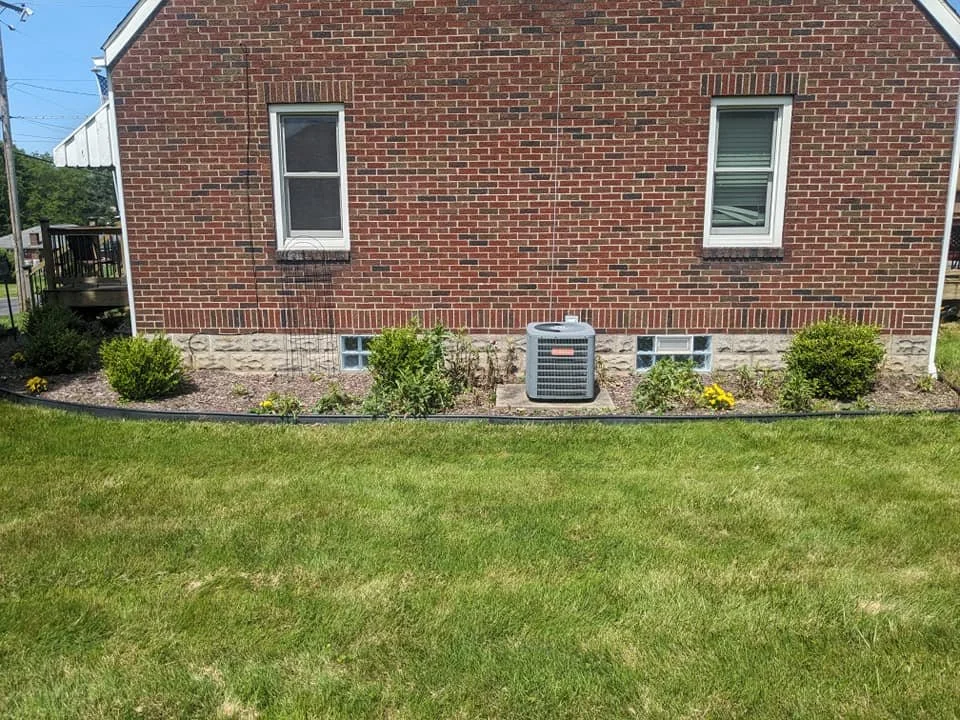 A brick house with two windows, a small garden bed with bushes and flowers, and an air conditioning unit on a small concrete pad on the ground. A green lawn is in the foreground.