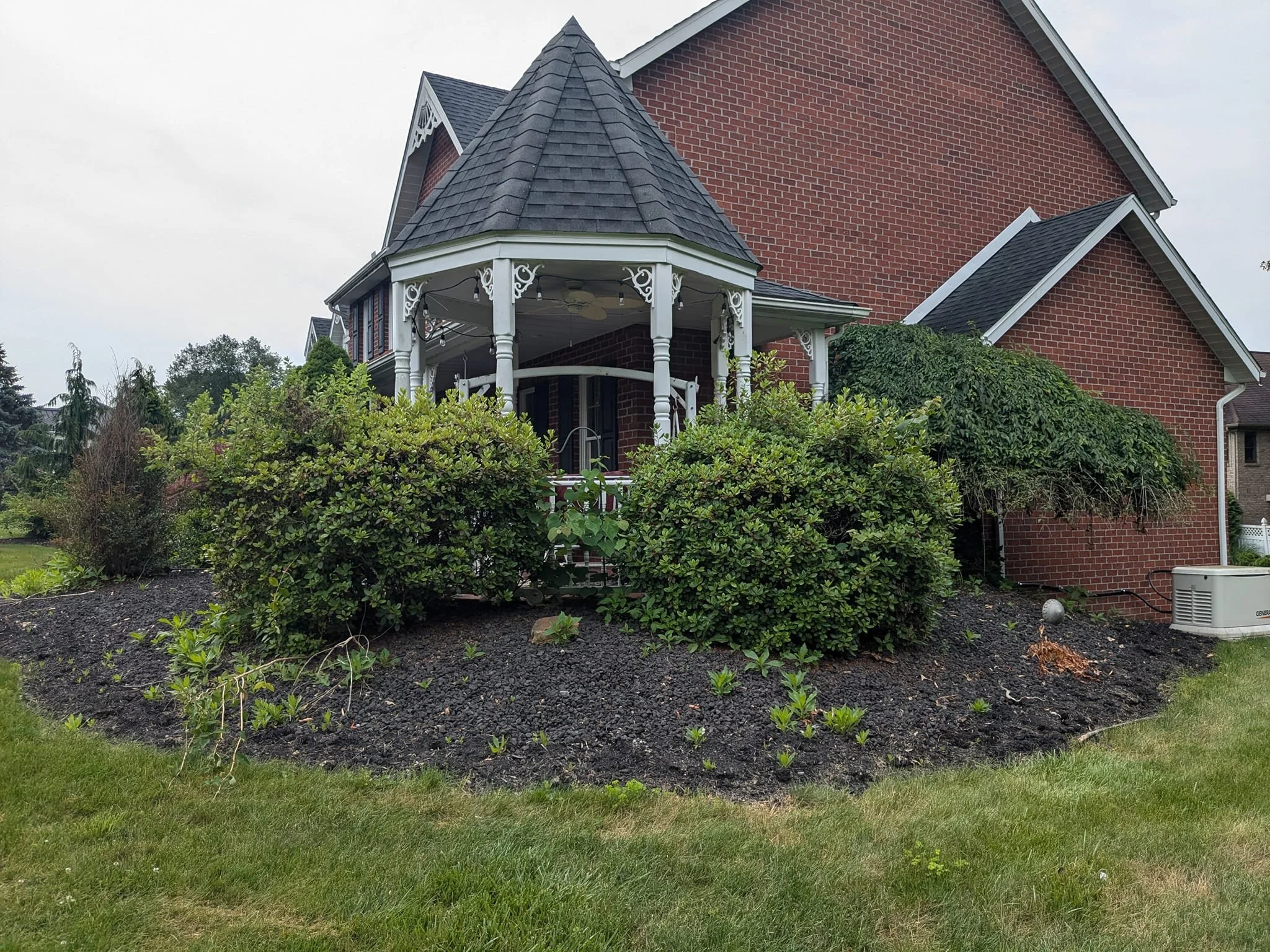 Brick house with a front porch, surrounded by green bushes and a flower bed with small plants, under an overcast sky.
