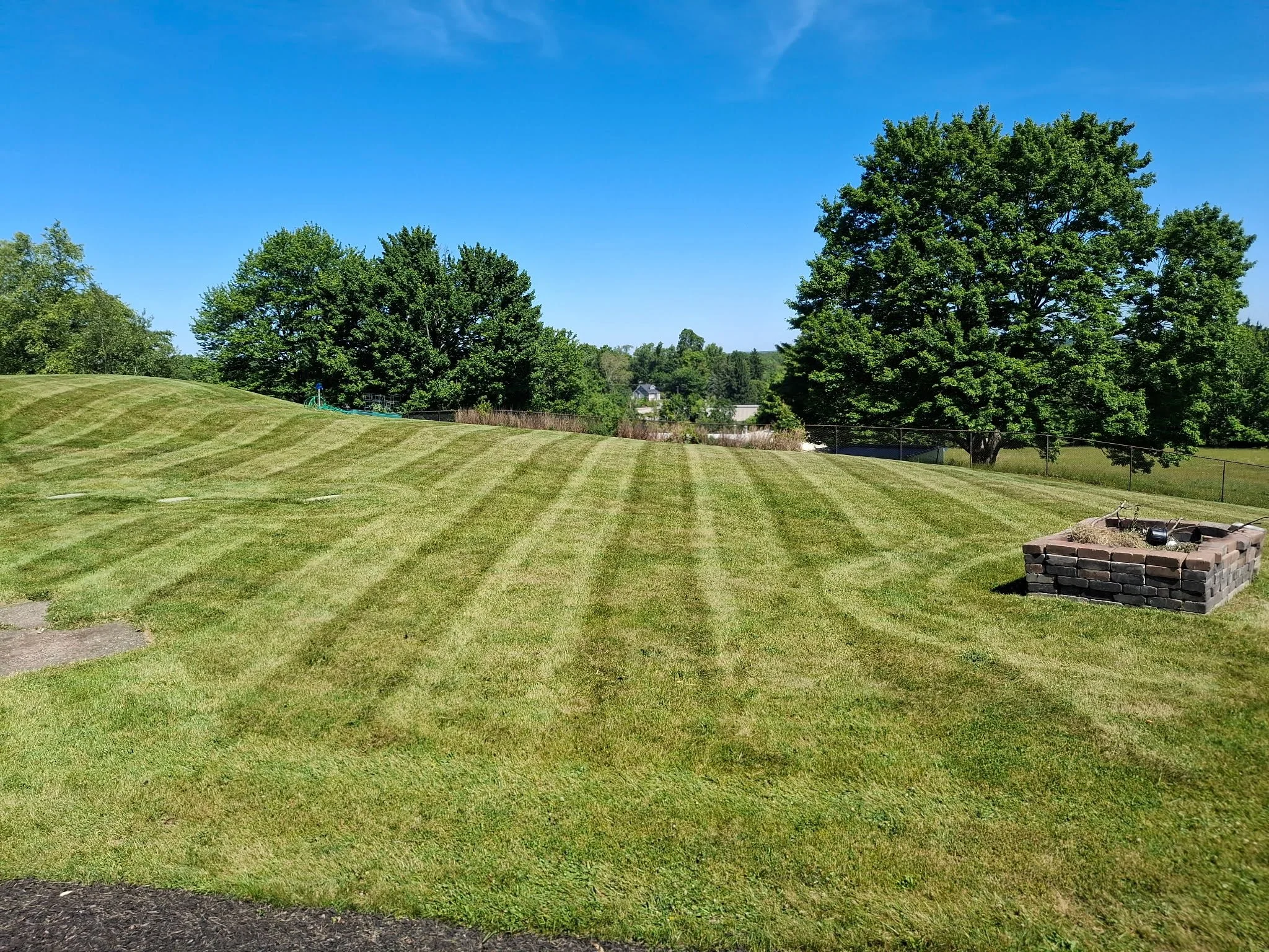A well-maintained grassy lawn with alternating light and dark green stripes, large trees in the background, a clear blue sky, and a small brick structure with garden tools in the foreground.