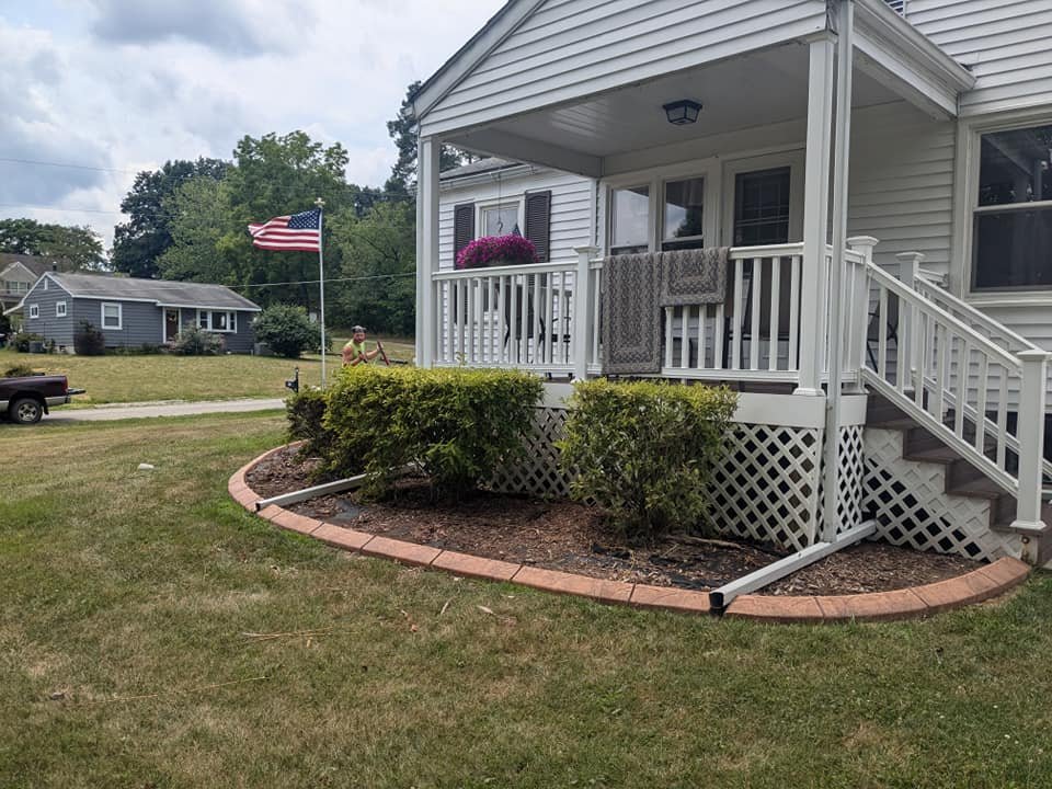 Front porch of a white house with stairs, a hanging flower basket, towels, and an American flag on a pole in the yard. A woman stands near the flag, and a dark-colored pickup truck is parked nearby.