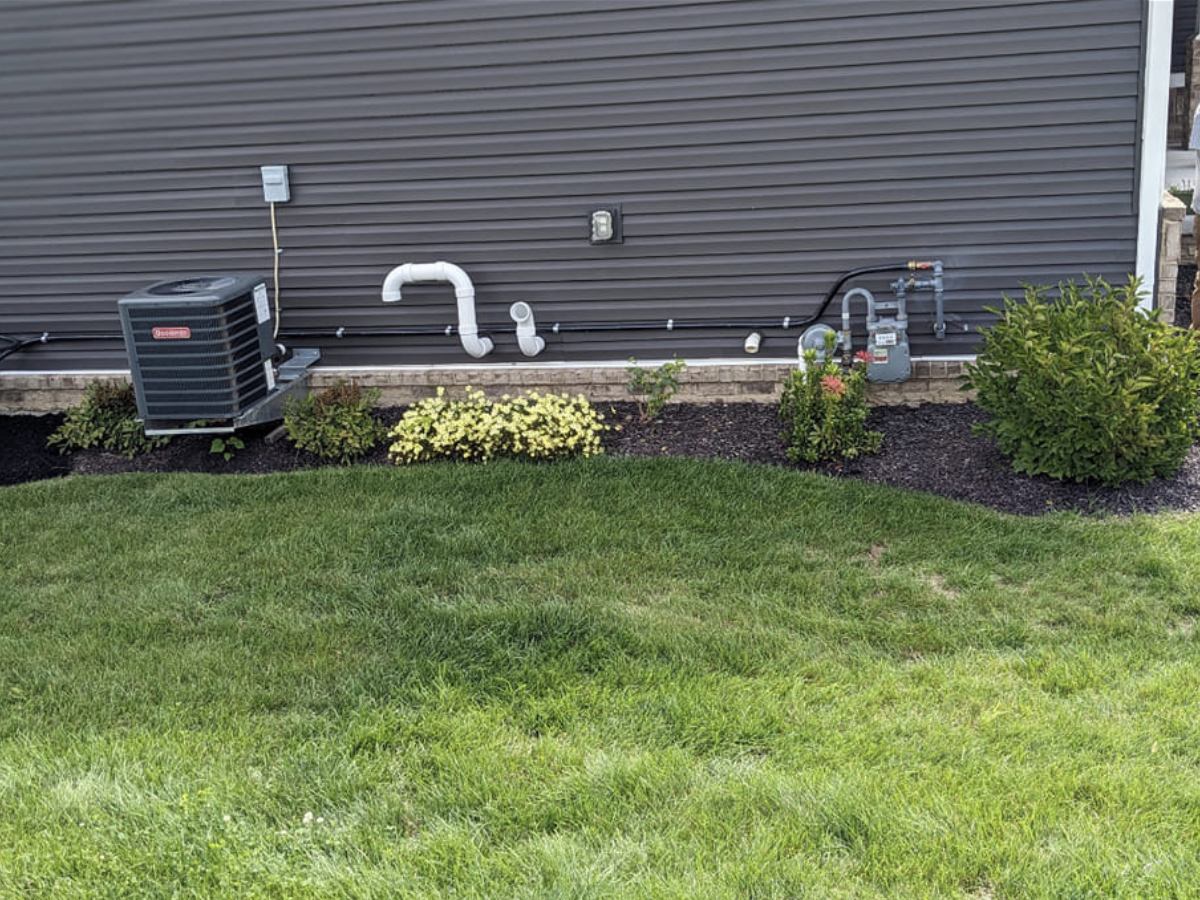 View of the side of a house with an HVAC unit, a white pipe, and green bushes in front of gray siding.