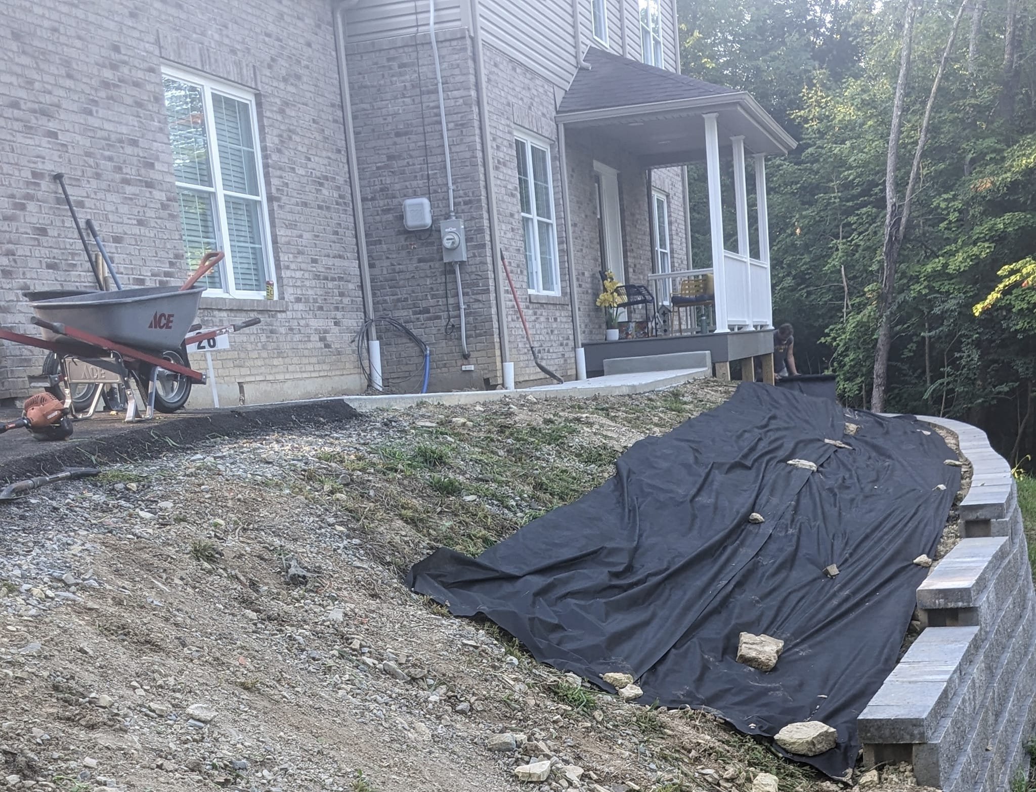 Backyard construction site with brick house, wheelbarrow, tools, mulch, and trees.