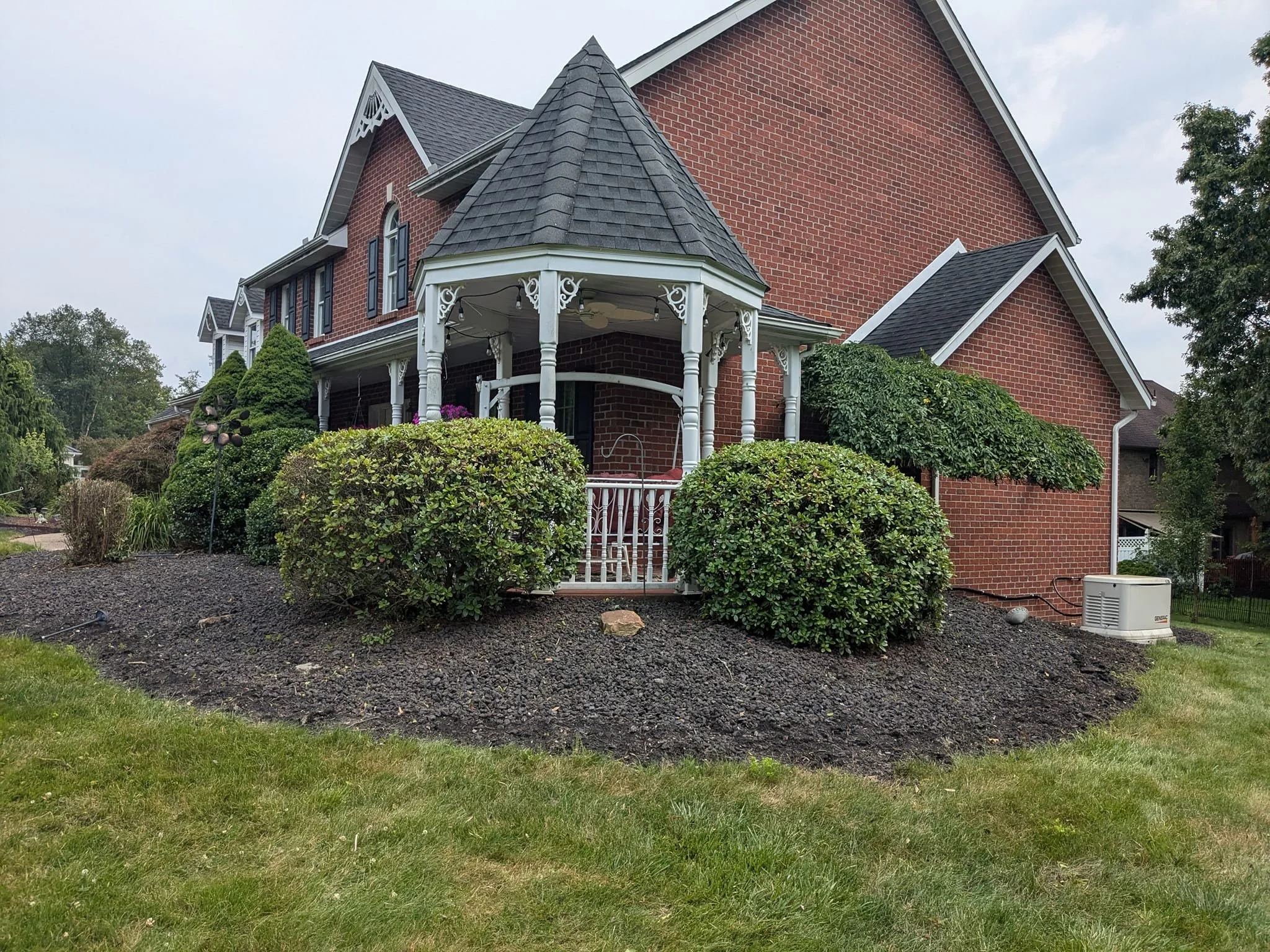 A large brick house with a pointed roof and a rounded turret with a conical roof, white trim, and decorative accents. The front yard features neatly trimmed bushes, small trees, and a landscaped garden with mulch and a pathway. The sky is overcast.