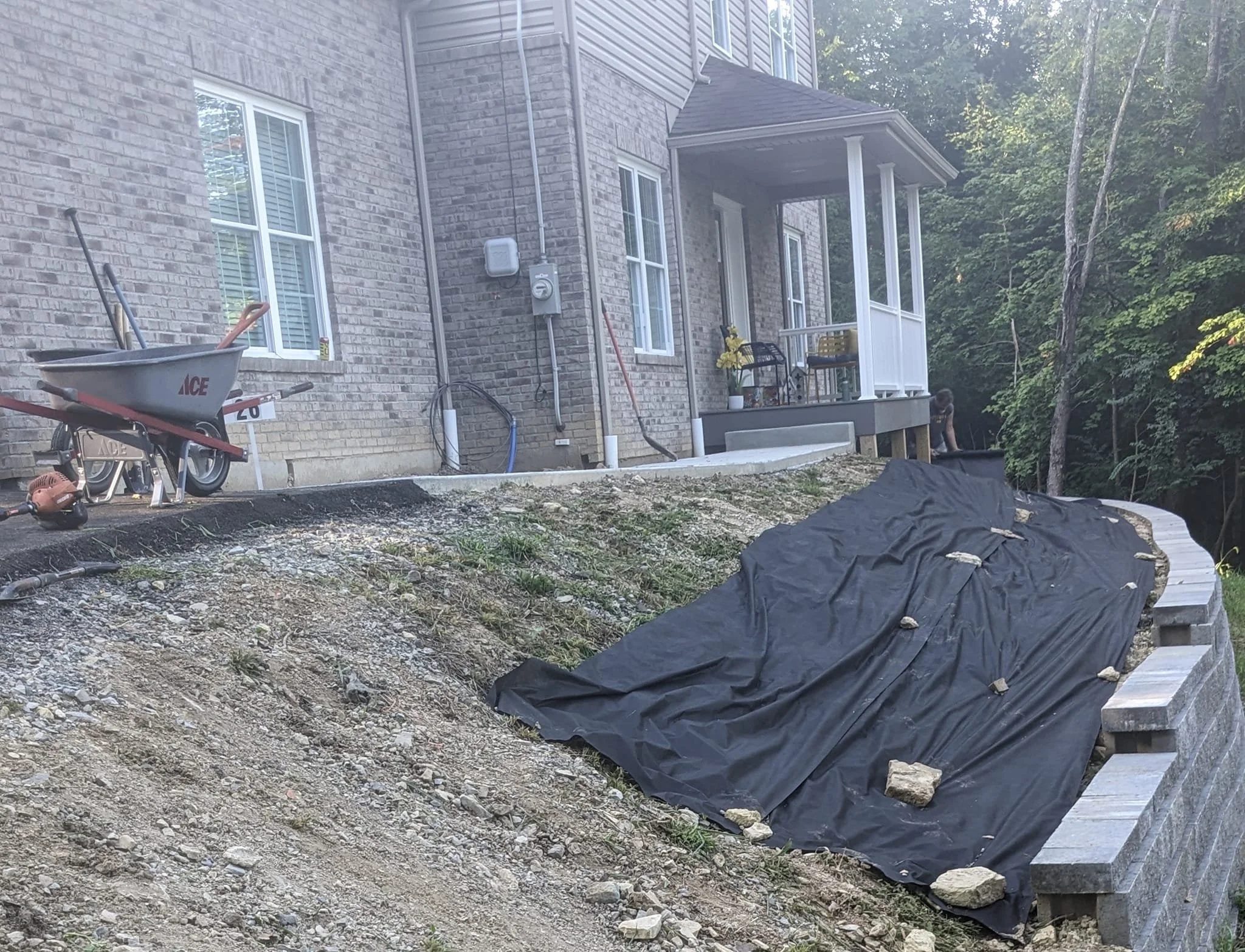 Construction site with a brick house, wheelbarrow, tools, black tarp, and construction materials on uneven ground.
