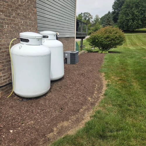 Two large propane tanks outside a house on a bed of mulch, with a lawn and trees in the background.