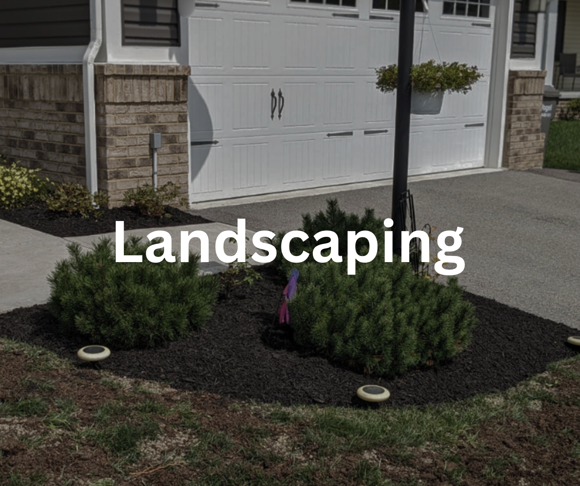 A landscaped front yard with small shrubs, mulch, and landscape lighting near a sidewalk. The house has a brick and siding facade with a white garage door, a hanging flower basket, and a black pole.