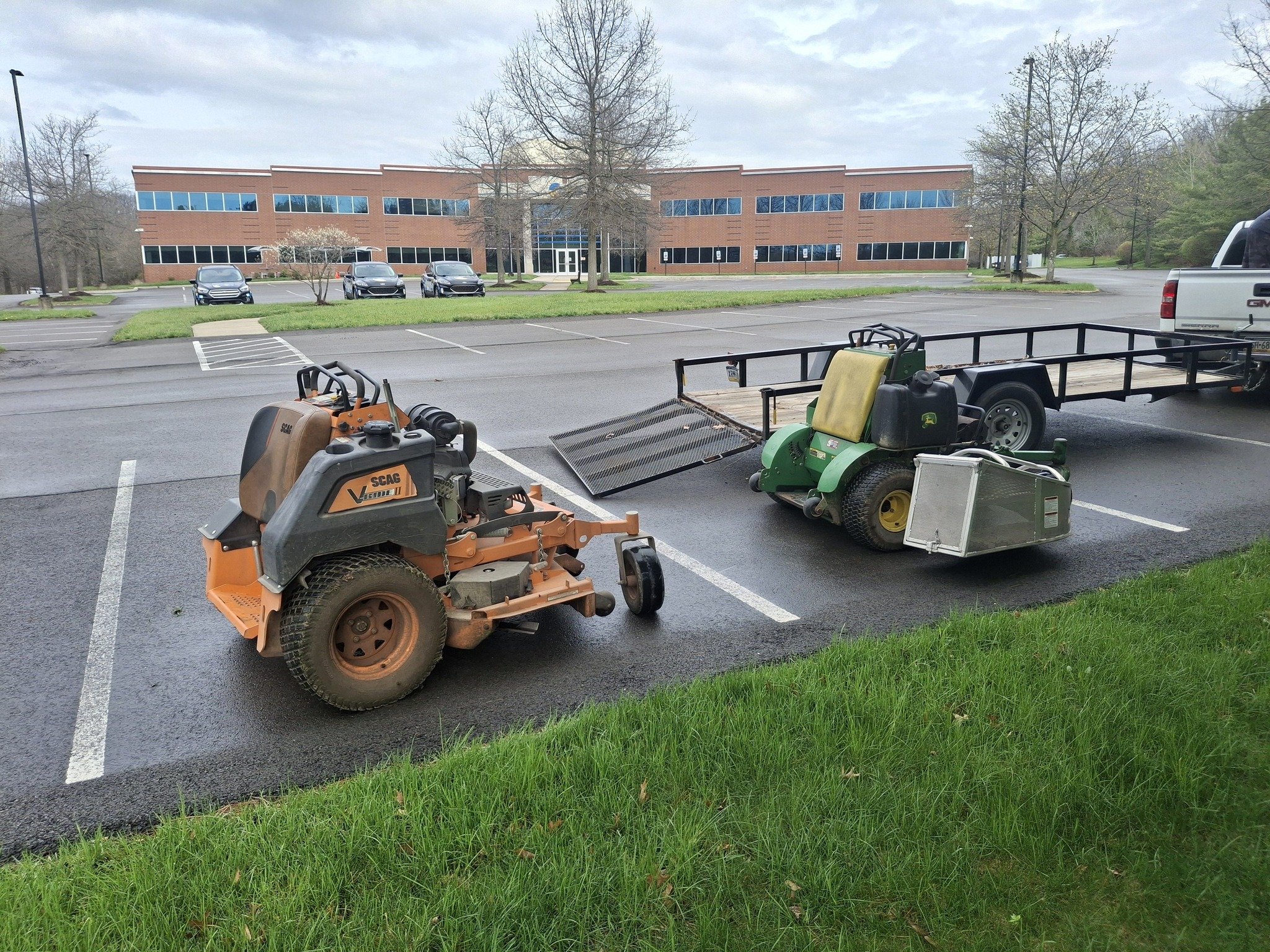 A parking lot with three small cars parked in front of a modern brick office building with multiple windows. The parking lot has some equipment trailer and lawn equipment in the foreground. Trees with no leaves are visible around the parking lot and 