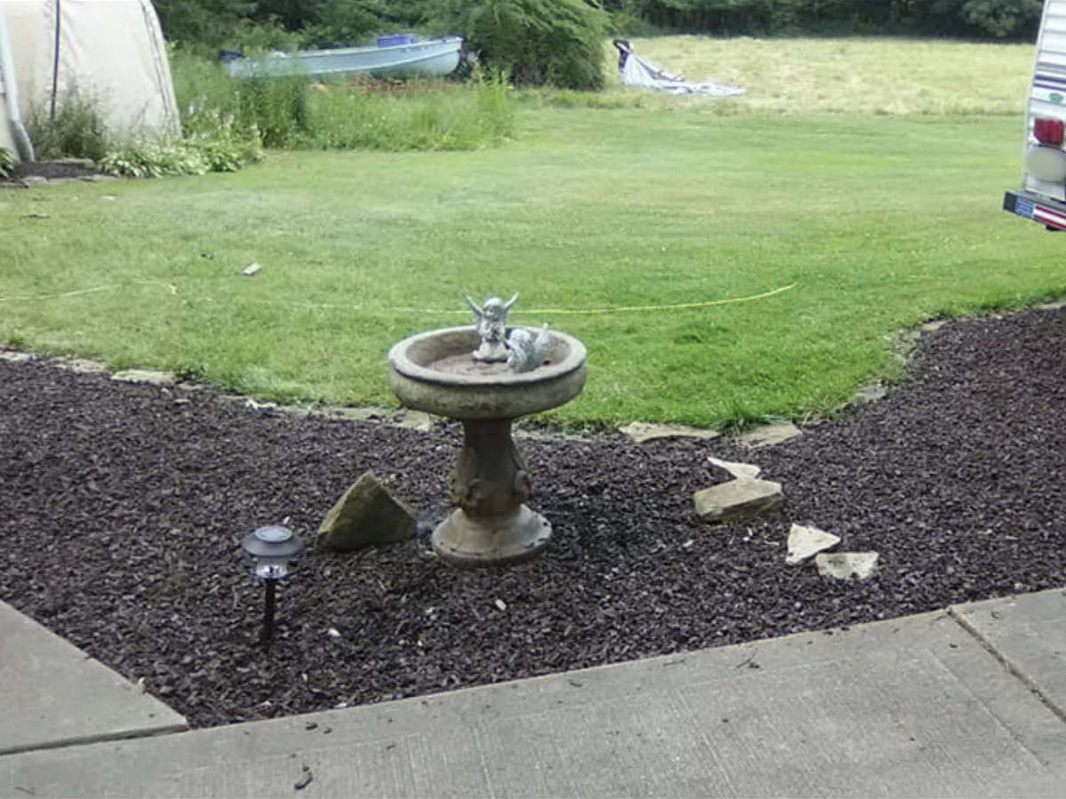 A garden scene with a bird bath containing a small decorative animal figure, surrounded by bark mulch and stepping stones, with a grassy yard and boats in the background.