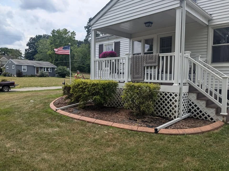 Front yard of a house with a porch, an American flag, bushes, and a person in the background.