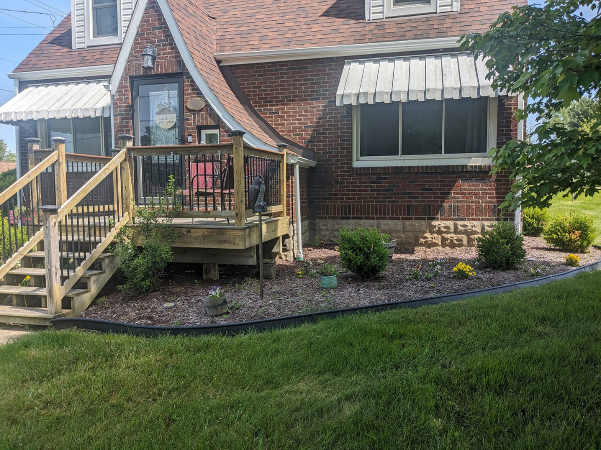 The image shows the exterior of a brick house with a small front yard garden that has flowering plants and bushes. There is a wooden deck with railings and steps leading up to a glass door with a sign that says 'little pumpkin.' A chair is visible on