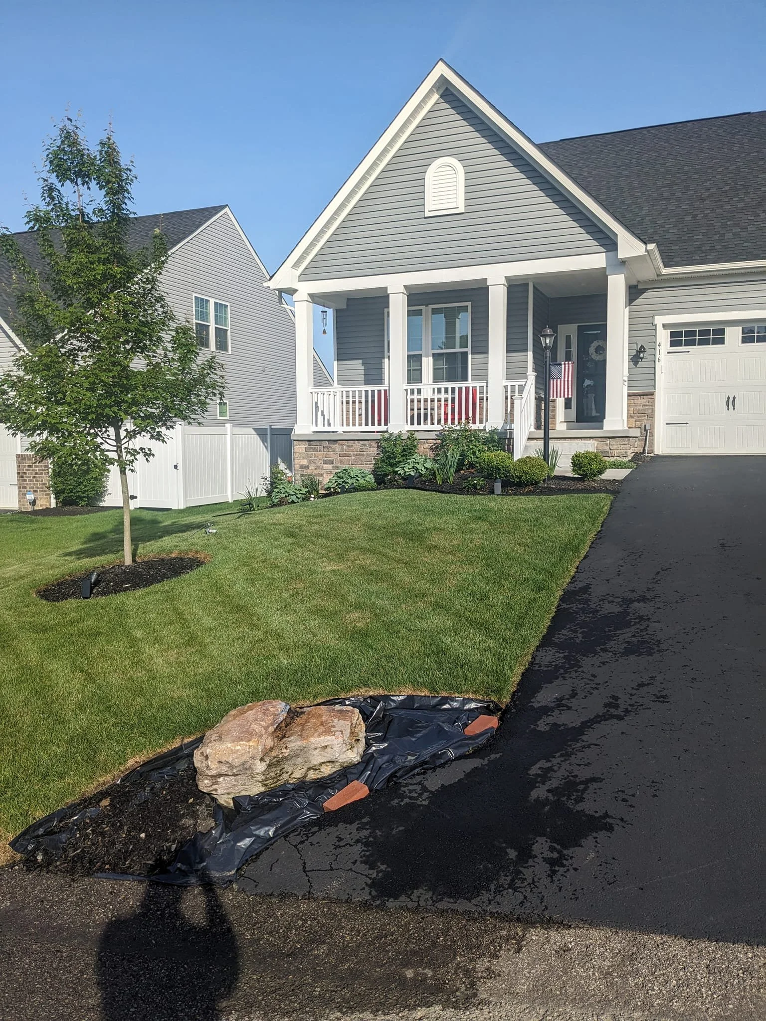 A newly planted small tree with mulch around the base and a fence in front of a house with gray siding and a porch, beside a black paved driveway, with a large rock, black plastic sheet, and small bricks on the ground.