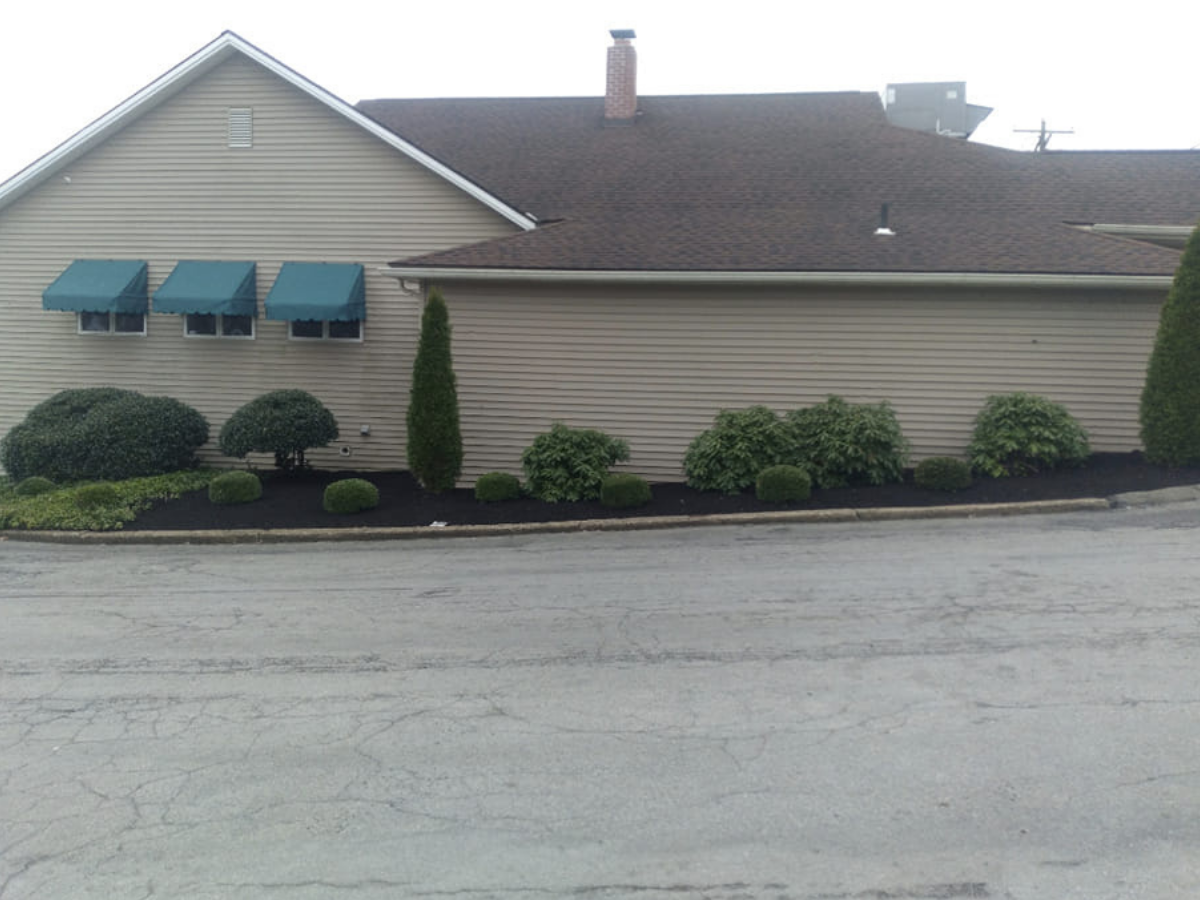 A beige residential building with three blue window awnings and well-manicured bushes and shrubs along the foundation, with an asphalt road in the foreground.