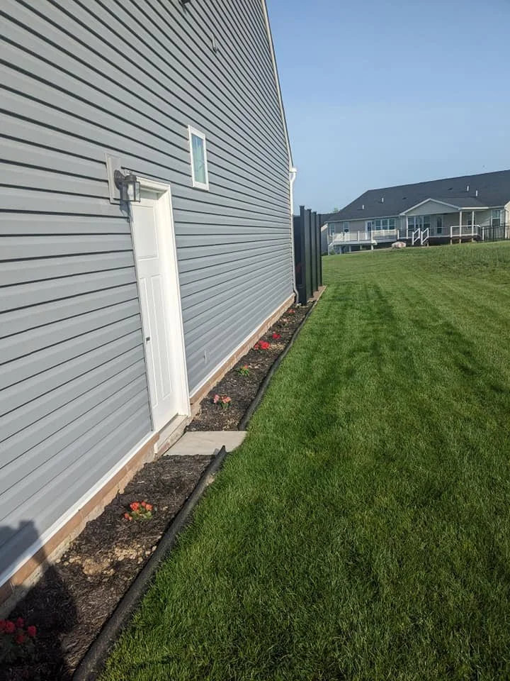 Side yard of a house with gray vinyl siding, a white door, and small flower bed with mulch and flowers along the foundation. There is a lawn and a neighboring house with a deck in the background.