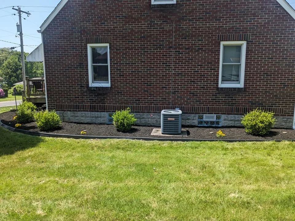 A brick house with two windows, a central air conditioning unit, and a landscaped area with small green bushes and flowers, bordered by a black edging along the yard.