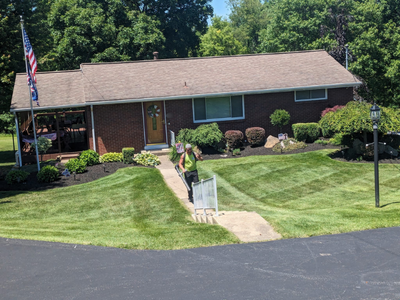 A woman walking on a sidewalk in front of a suburban brick house with a well-maintained lawn, American flag, and street lamp.