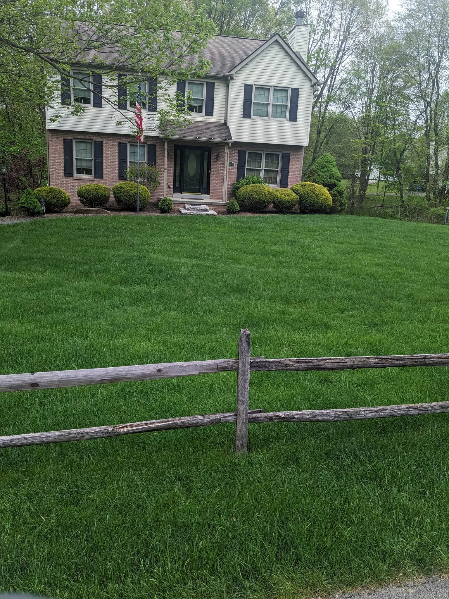 A two-story house with a brick lower level and white upper siding, surrounded by a well-maintained lawn and bushes, with a wooden fence in the foreground and trees in the background.