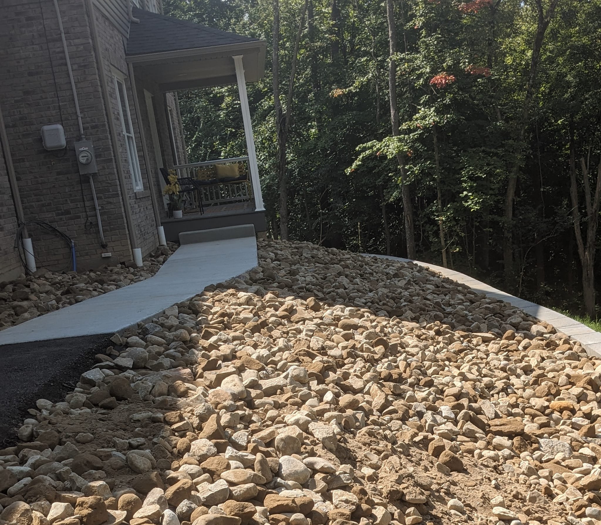 A house with a small porch and a black chair, surrounded by trees, with a pathway made of large rocks leading up to the porch.