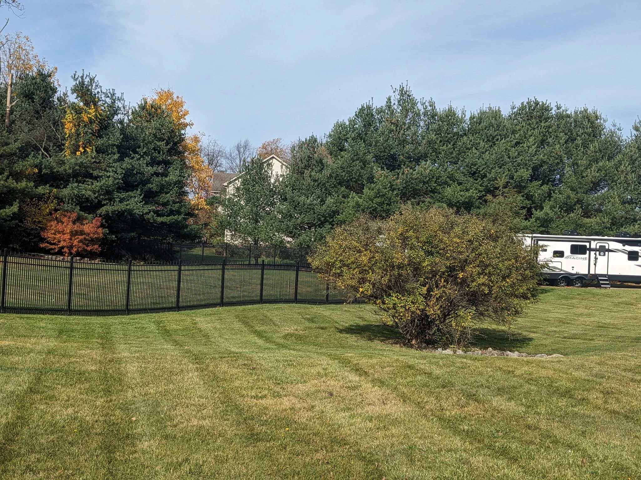 A suburban backyard with a lush green lawn, a black metal fence, and various trees, including some with autumn-colored leaves. In the background, a white house and a parked recreational vehicle (RV) are visible under a blue sky with some clouds.