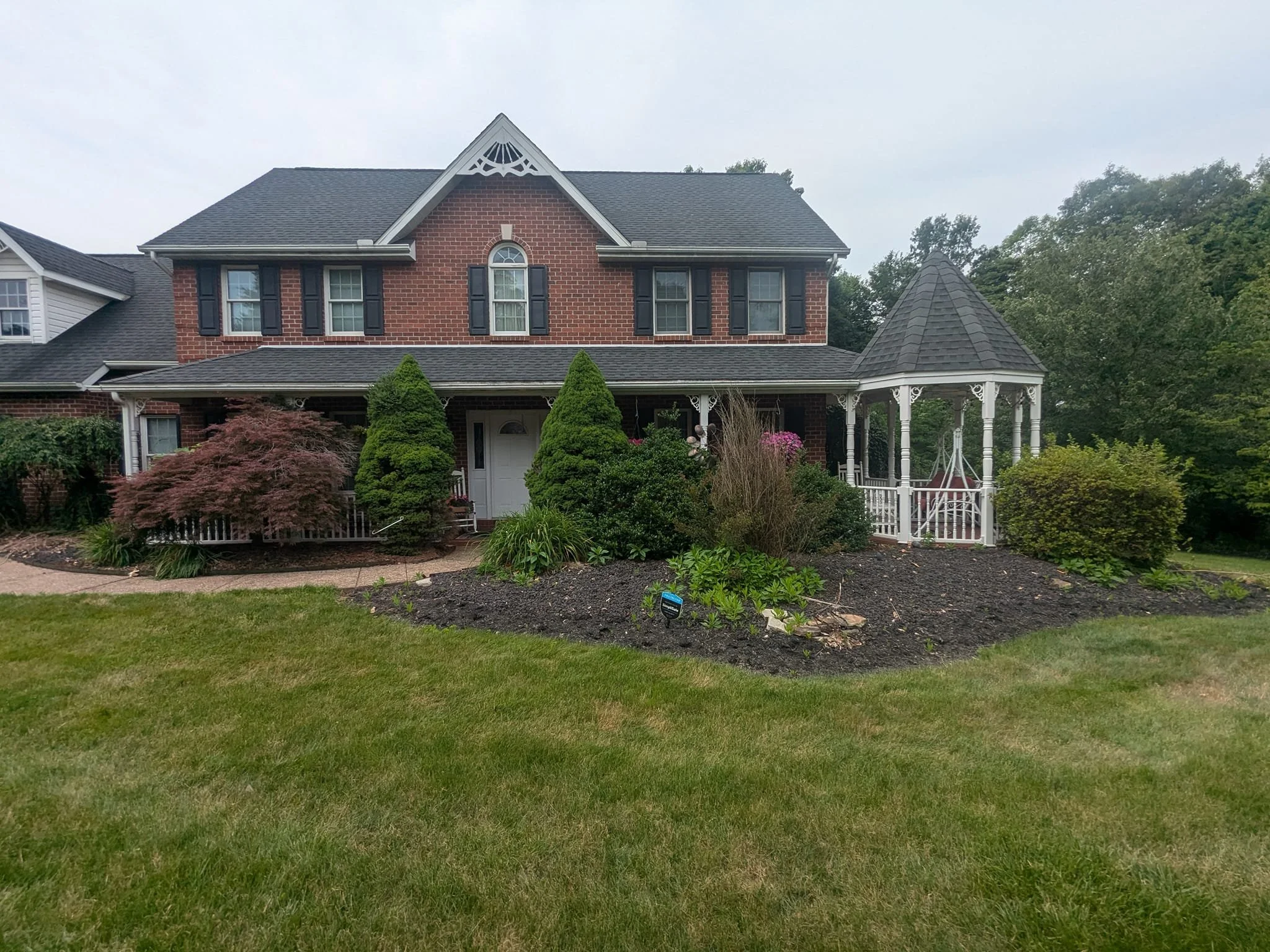 A two-story brick house with black shutters, a front porch with white decorative trim, and a flower garden in front. The house has a gabled roof with an ornate triangular detail and a conical turret on the right side. There are lush green trees in th