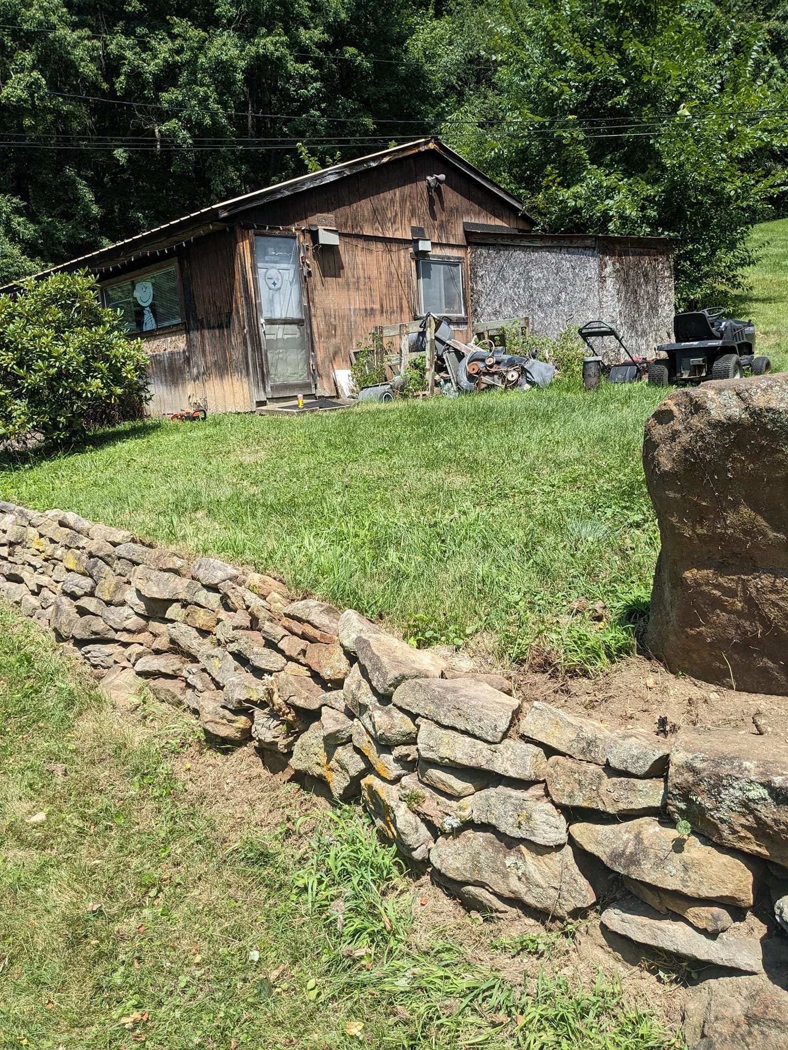 A small wooden shed with a sloped roof, surrounded by grassy lawn and trees. There are various discarded items like a lawn mower, motorcycle parts, and a toy in front of the shed. A stone wall is in the foreground.