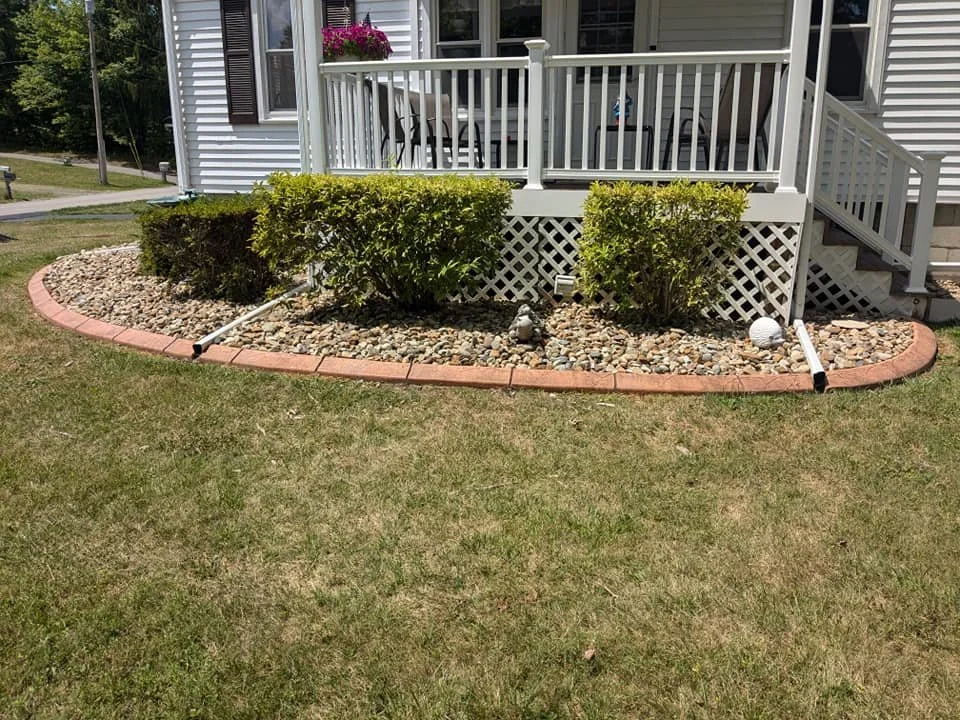 A landscaped front yard with a small garden bed edged with red bricks, containing green bushes, small decorative statues, and a white spherical garden ornament. The garden bed is covered with rocks, and there are white drainage pipes on each side of 