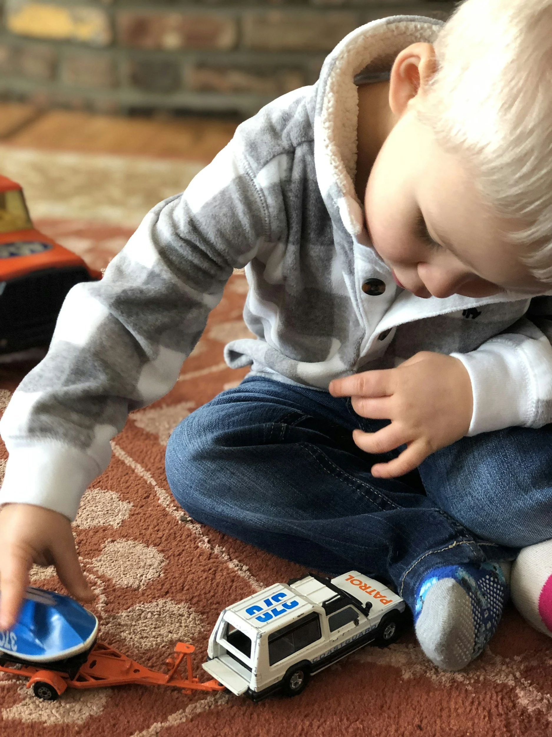 A child with blonde hair playing with toy rescue vehicles on a patterned carpet, with a brick wall in the background.